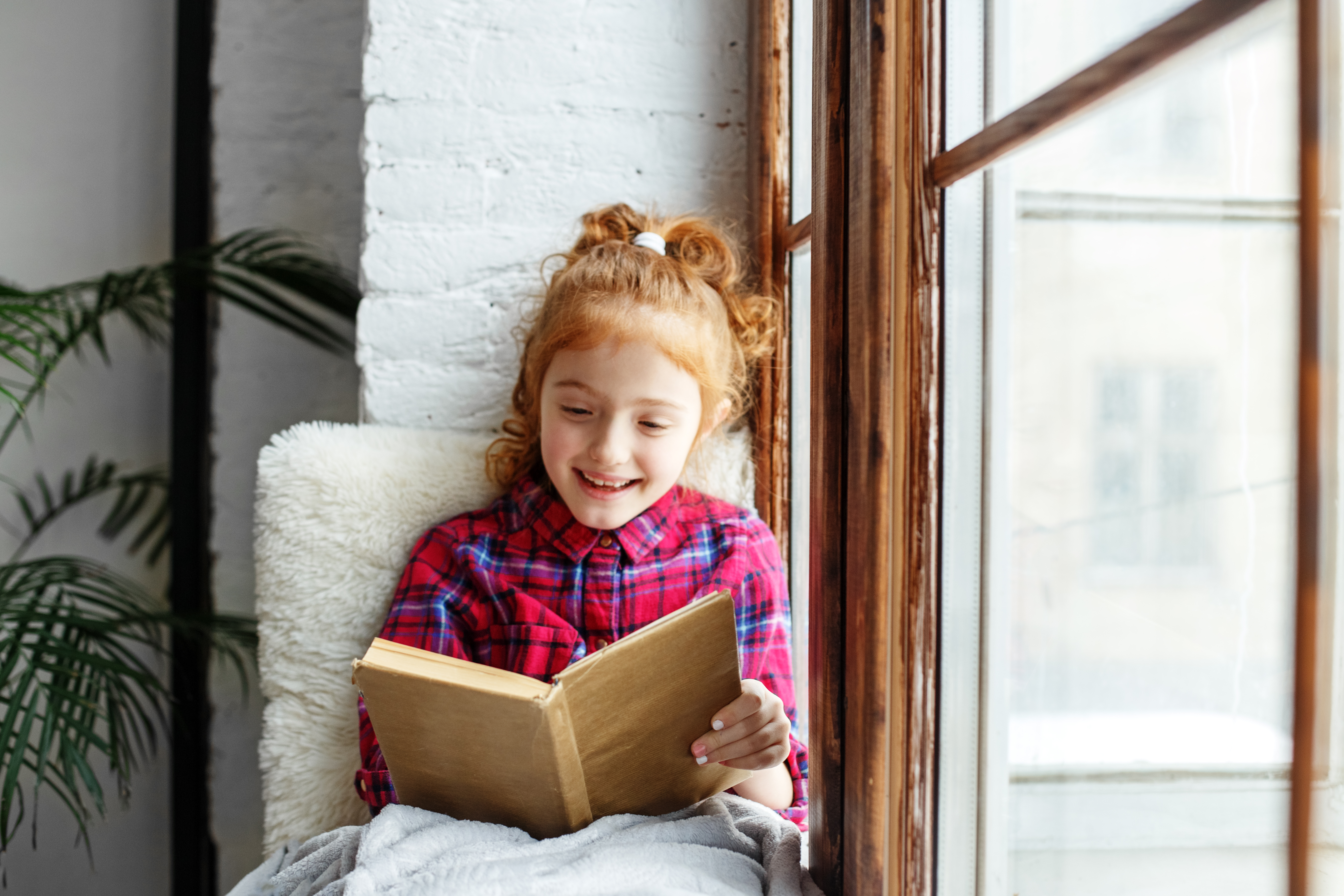 Smiling girl with red hair reads a book next to a sunny window.